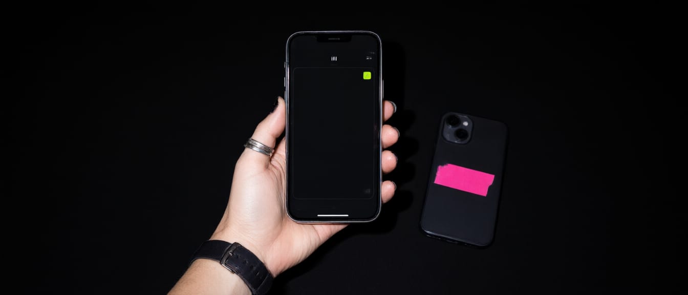 Flash photo: a ringed hand holding a phone showing a launch card, a second phone face down under pink gaffer tape